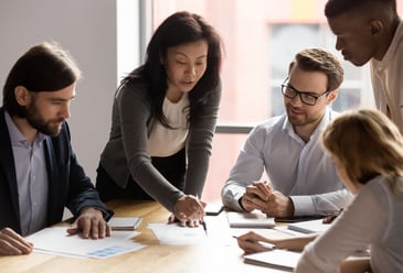 A manager managing four people on her team with a Coaching leadership style as they sit as a table outlining the data on pieces of paper in front of them. 