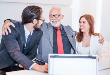 A brother and sister sitting either side of their Dad who has his arm around each of them as they smile over an achievement within their family business that they manage together. 