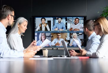 Four people sitting in a board room during a meeting with six members of their team on a screen who are hybrid working therefore attending the meeting virtually which is the new normal since the pandemic. 