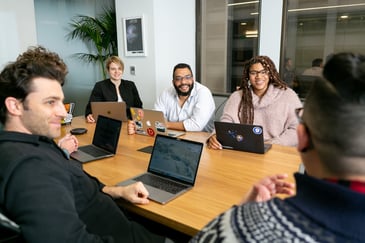 Five managers sitting around the table in a meeting room with laptops open in front of them as they have a productive and effective leadership team meeting. 