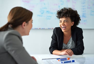 Two women sitting at a table in front of a whiteboard at work laughing as they have an optimistic and productive meeting together. 
