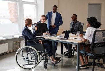 A diverse team including one team member in a wheelchair, three people of colour, and a white male, having a meeting in the workplace without any judgement or lack trust in the other's knowledge and skills. 