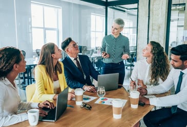 A first time manager standing over a table where five team members sit listening intently and taking notes in notebooks and on their laptops. 