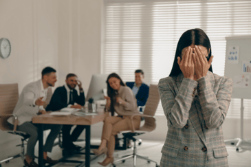 Four smiling employees in an office, sitting at a desk in a conversation whilst another employee is hiding behind her hands struggling with emotional safety. 