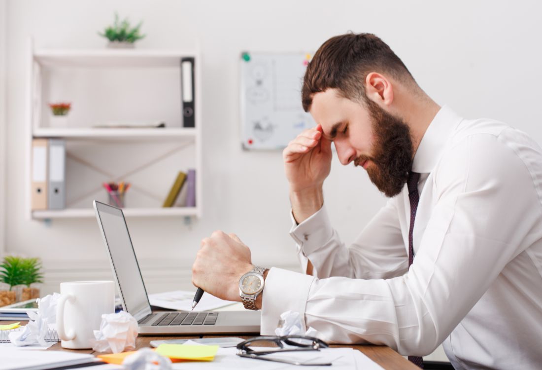 A tired businessman leaning over his desk with his head in his hands and laptop in front of him as he struggles to cope with a difficult situation.