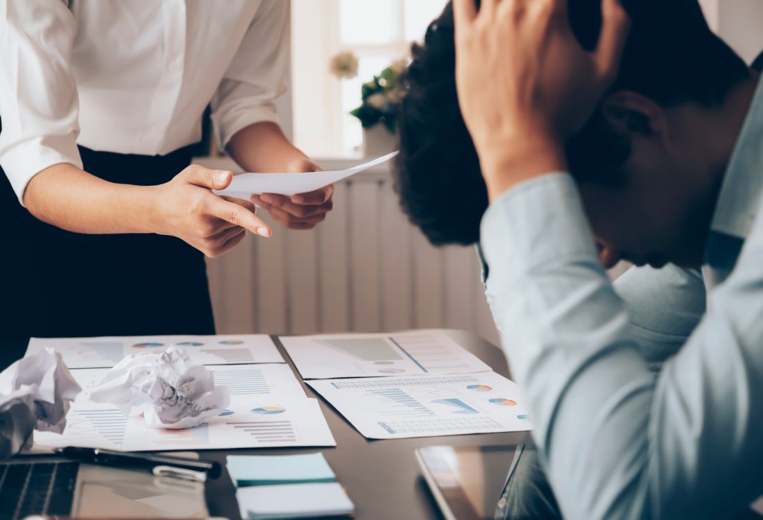 A coworker leaning over another berating him and pointing her finger accusingly with a piece of paper in her hands as she writes him off for being difficult instead of listening to him and his side of the story.