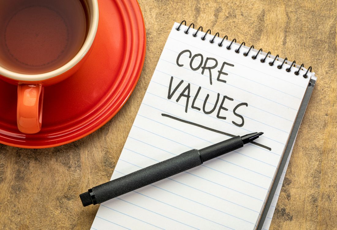 The words Core Values with an underline under the word Values written on a notebook next to a cup of tea and a pen on the notebook waiting to be used by the business owner about to write out their business' values.