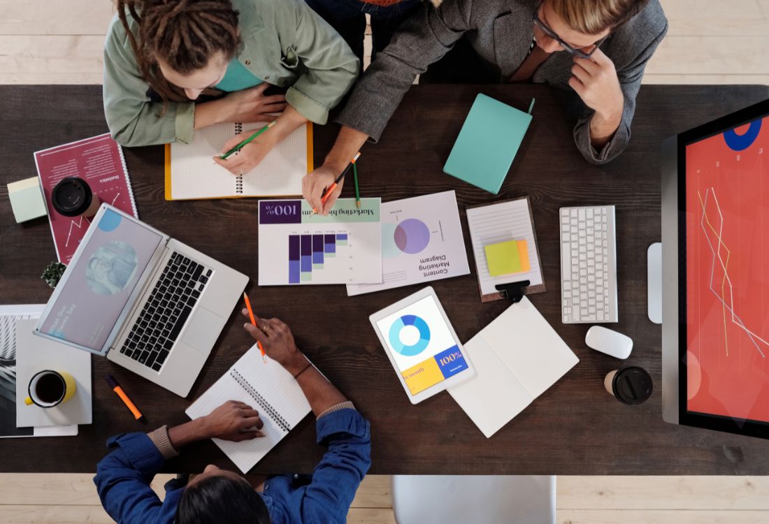 A team of three colleagues sitting around a table with their laptops, notebooks and notes from slides as they work collaboratively on a project for their workplace.