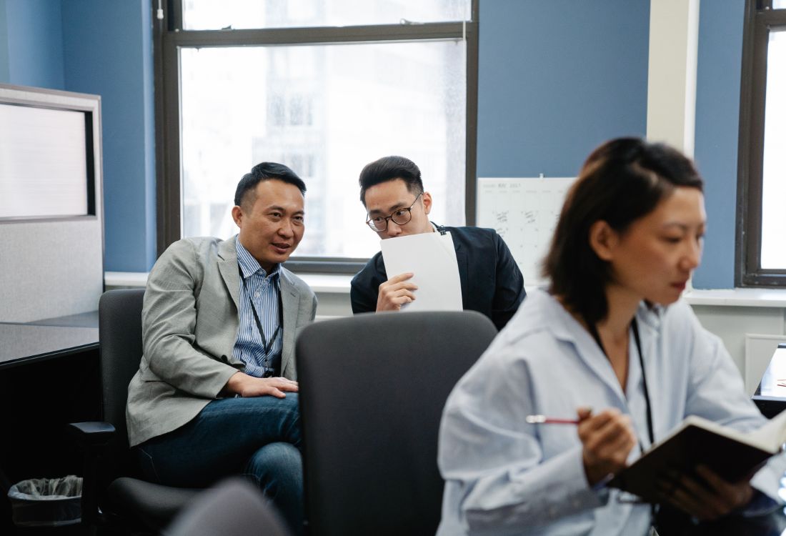 Two male colleagues gossiping at their desk looking over at the female colleague they are gossiping about who is trying to make notes at her desk.