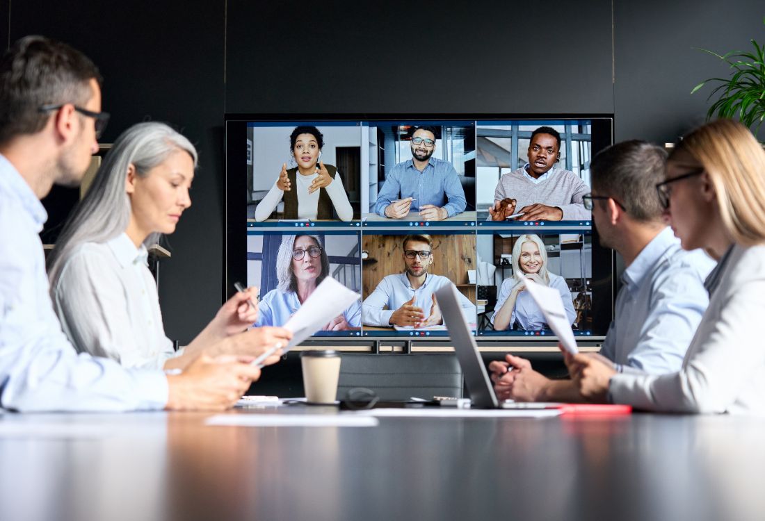 Four people sitting in a board room during a meeting with six members of their team on a screen who are hybrid working therefore attending the meeting virtually which is the new normal since the pandemic.