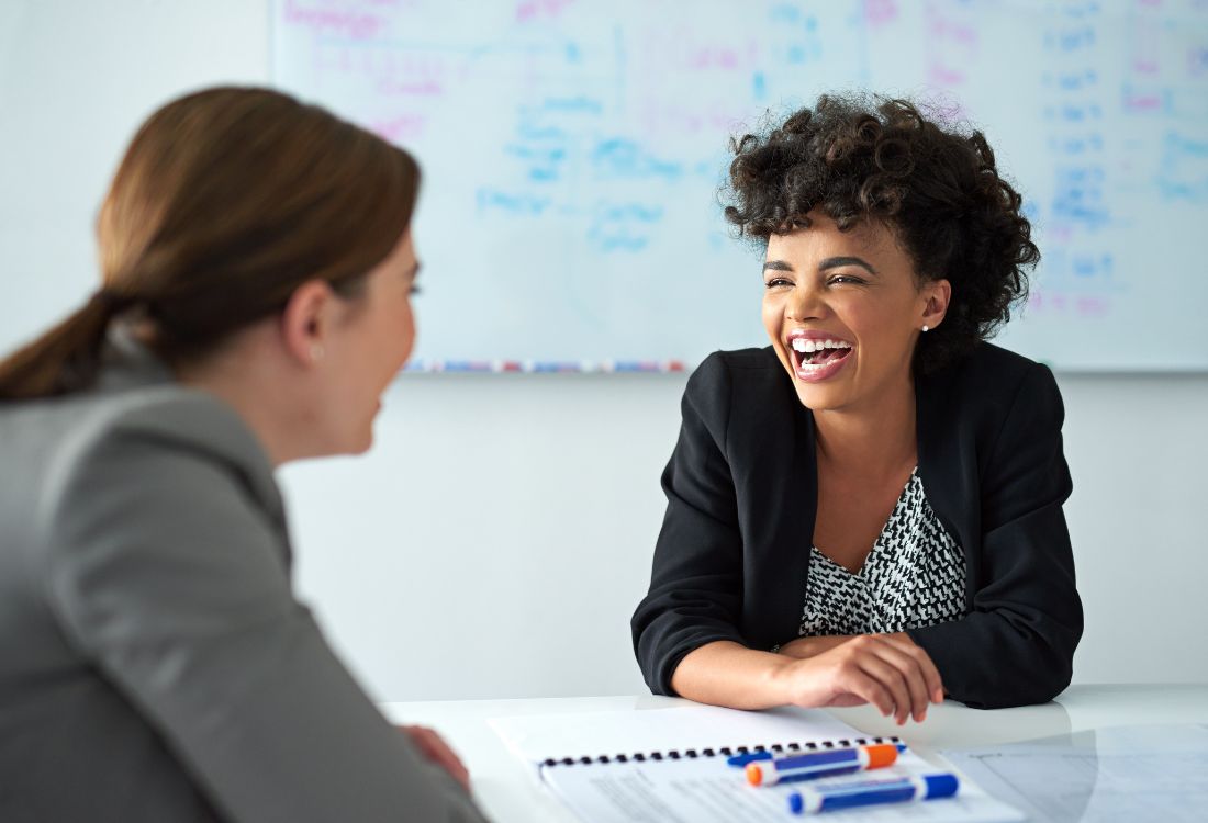 Two women sitting at a table in front of a whiteboard at work laughing as they have an optimistic and productive meeting together.