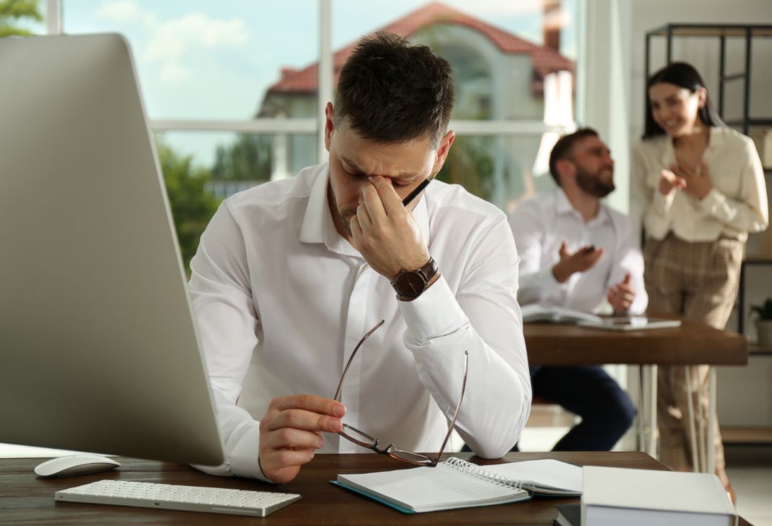 A man leaning over his desk in front of his PC with his head in his hands as he pinches his nose in frustration as two of his colleagues gossip behind him creating a toxic environment complemented by a toxic workplace culture.