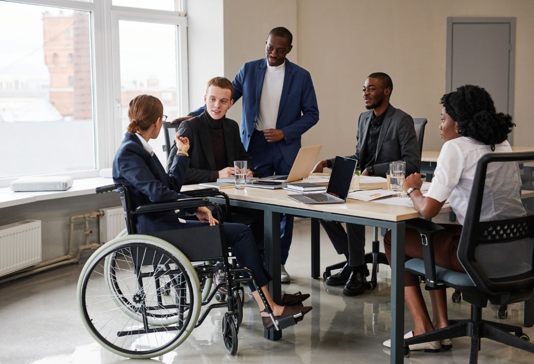 A diverse team including one team member in a wheelchair, three people of colour, and a white male, having a meeting in the workplace without any judgement or lack trust in the other's knowledge and skills.