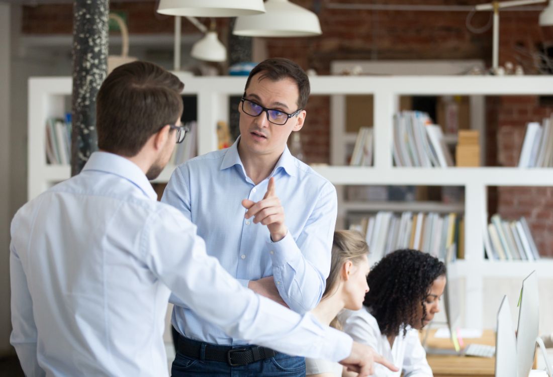 Two male colleagues having a crucial conversation an office with two woman working behind them trying to work as the two men sort their differences even though one of them has not checked his motives for the conversation.