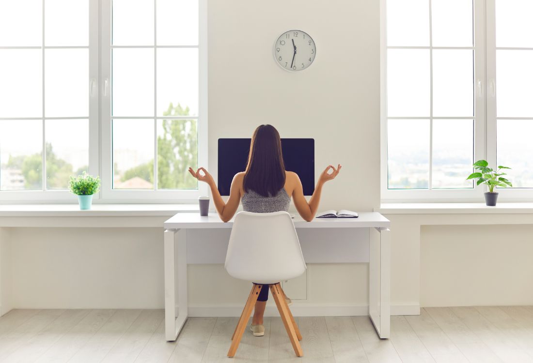 A business woman sitting at her desk in her clean and minimalist office meditating as she improves her workplace wellbeing by taking small breaks during the work day.