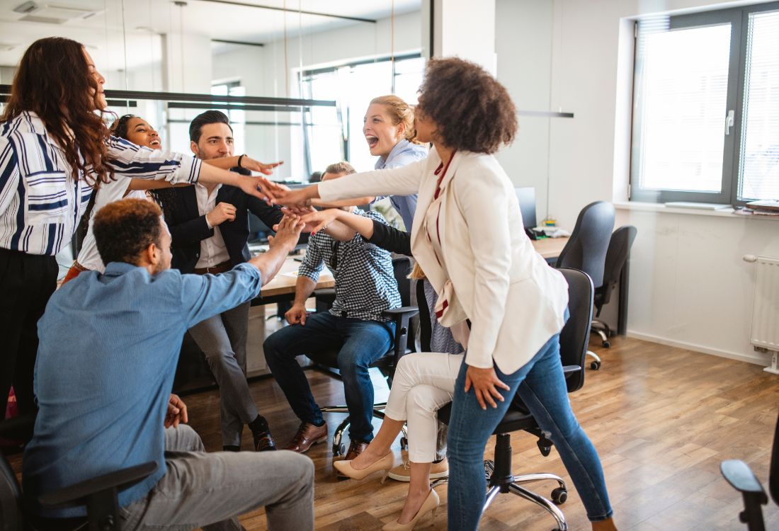 Seven colleagues laughing and smiling together as they put their hands on top of one another after a successful experiential learning session to help bring a positive behaviour change to their workplace.