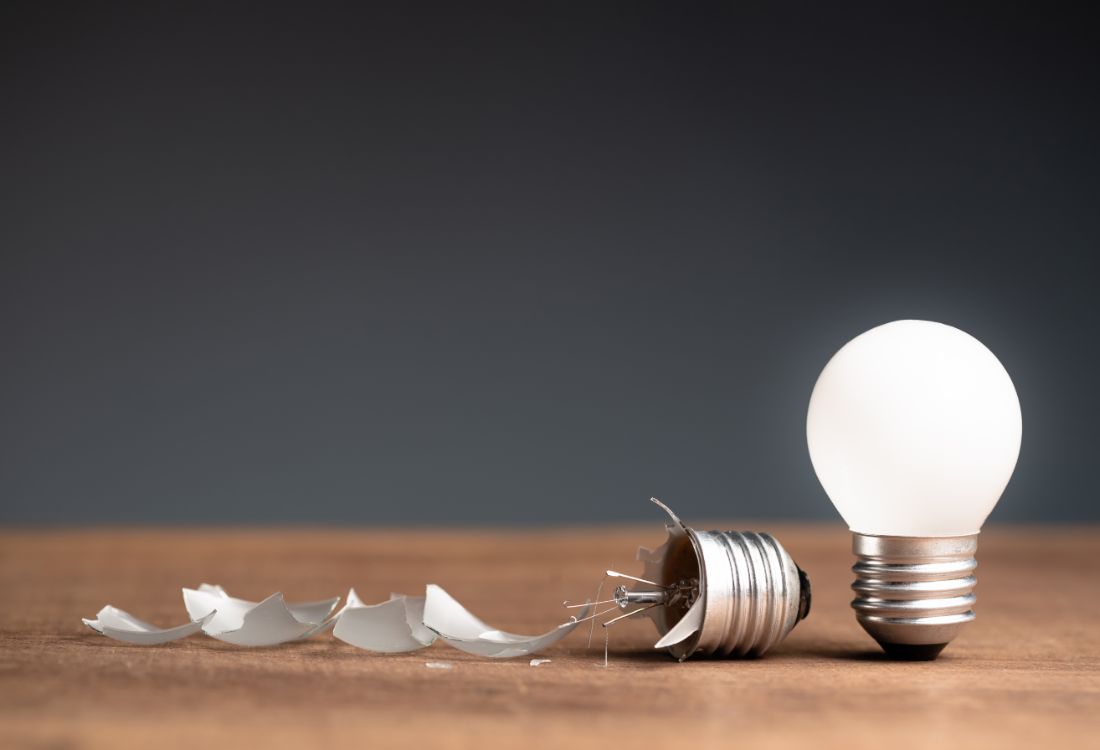 Two lightbulbs standing next to each other with the righthand one shining brightly and the left hand one smashed and broken on the ground as it has failed to shine.