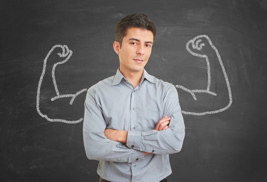 A businessman standing in front of a blackboard with a set of muscly arms drawn on it in white chalk, about to discuss the differences between confidence and arrogance. 