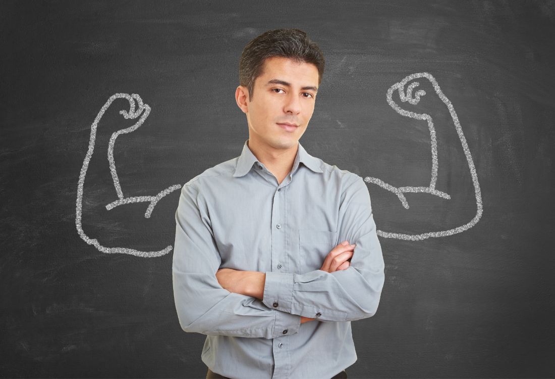 A businessman standing in front of a blackboard with a set of muscly arms drawn on it in white chalk, about to discuss the differences between confidence and arrogance.  