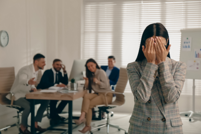 Four smiling employees in an office, sitting at a desk in a conversation whilst another employee is hiding behind her hands struggling with emotional safety.